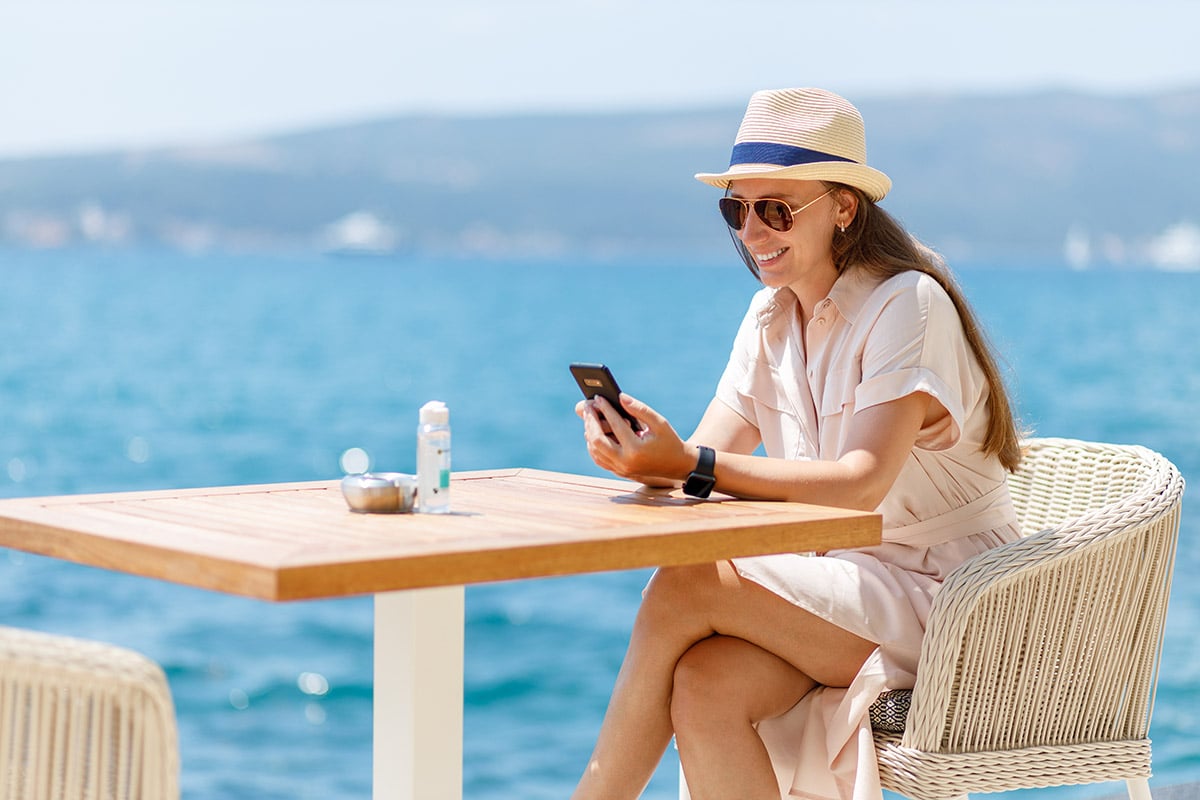 A traveler relaxes by the waterfront, checking her phone for new, creative rewards from her travel loyalty program.